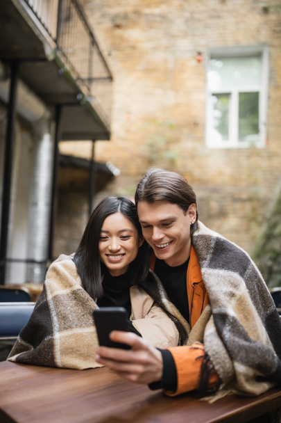 Couple interracial positif dans la couverture en utilisant le téléphone portable sur la terrasse du café  - Photo, image