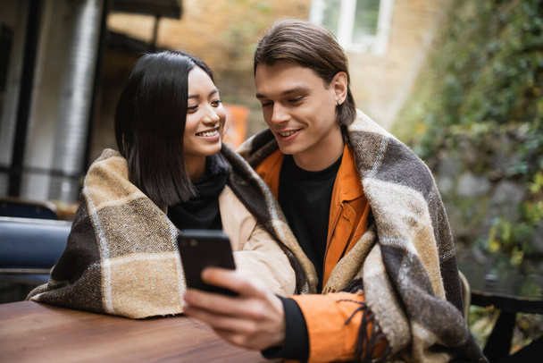 Mujer asiática sonriente en manta mirando novio usando teléfono inteligente borroso en café al aire libre  - Foto, imagen