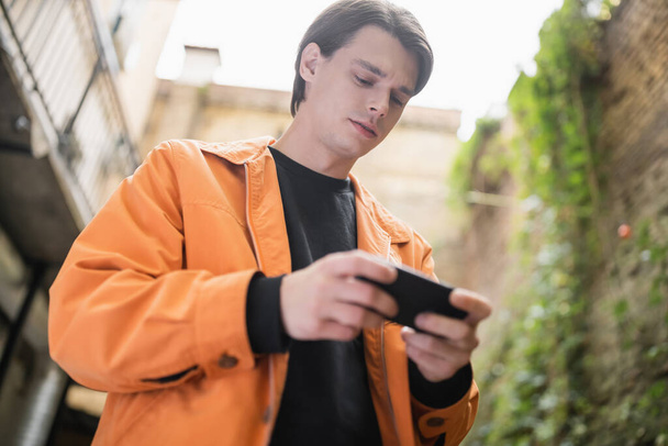 Низкий угол обзора молодого человека с помощью мобильного телефона на террасе кафе  - Фото, изображение