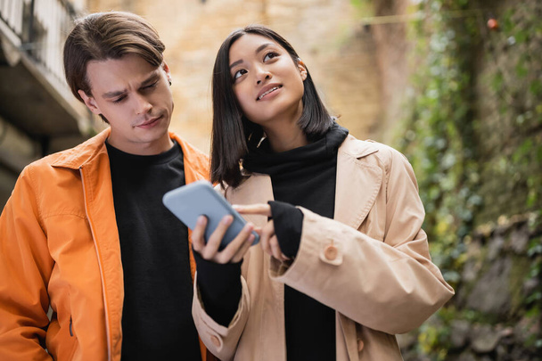 Low angle view of asian woman pointing at smartphone near boyfriend outdoors  - Photo, Image