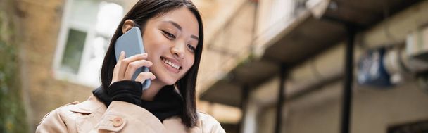 positive asiatique femme en trench coat parler sur smartphone à l'extérieur, bannière  - Photo, image