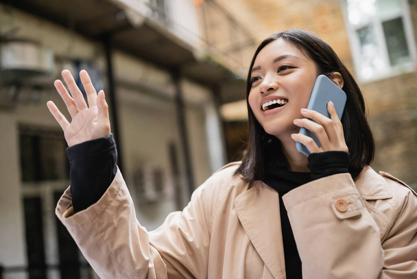 Sonriendo mujer asiática hablando en el teléfono inteligente y agitando la mano al aire libre  - Foto, Imagen