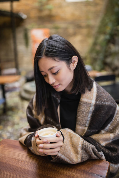 Joven mujer asiática en manta sosteniendo taza de papel con capuchino en café al aire libre  - Foto, imagen