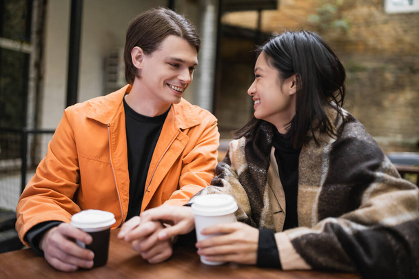 Couple interracial souriant avec couverture tenant la main près du café sur la terrasse du café  - Photo, image