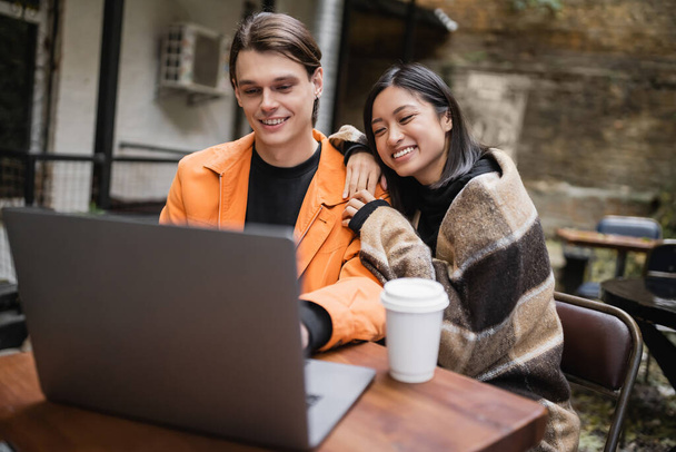 Smiling asian woman in blanket hugging boyfriend near laptop and coffee in outdoor cafe  - Foto, imagen