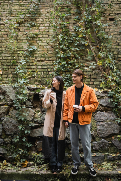 Νεαρό διαφυλετικό ζευγάρι κρατώντας καφέ για να πάει και κρατώντας τα χέρια κοντά στον τοίχο σε εξωτερικούς χώρους  - Φωτογραφία, εικόνα