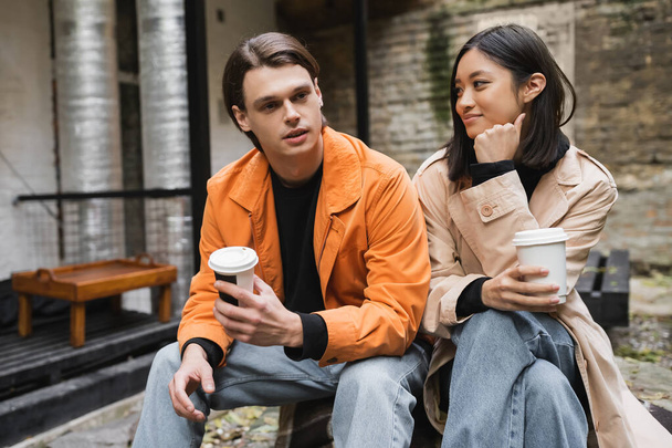 Young man in jacket holding coffee to go and talking to asian girlfriend on terrace of cafe  - Foto, imagen