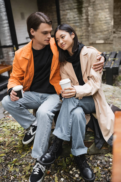 Young man hugging asian girlfriend in trench coat and holding takeaway drink in outdoor cafe  - Valokuva, kuva