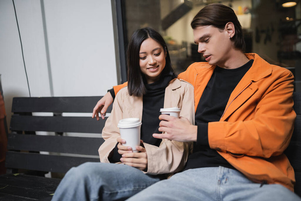 Joven pareja interracial sosteniendo café para ir mientras está sentado en el banco en la cafetería al aire libre - Foto, Imagen