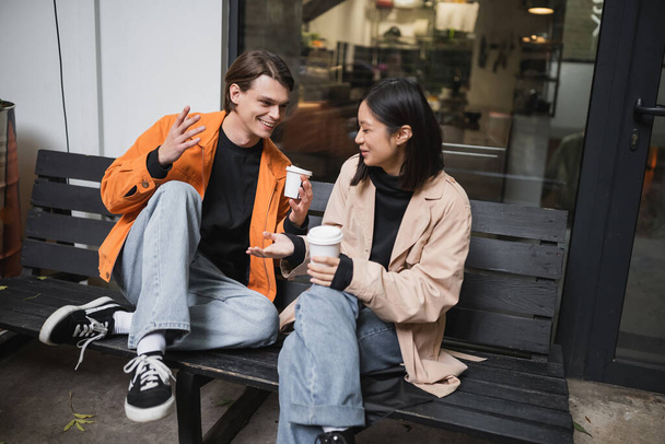 Young multiethnic couple holding paper cups while talking on bench near cafe  - Photo, Image