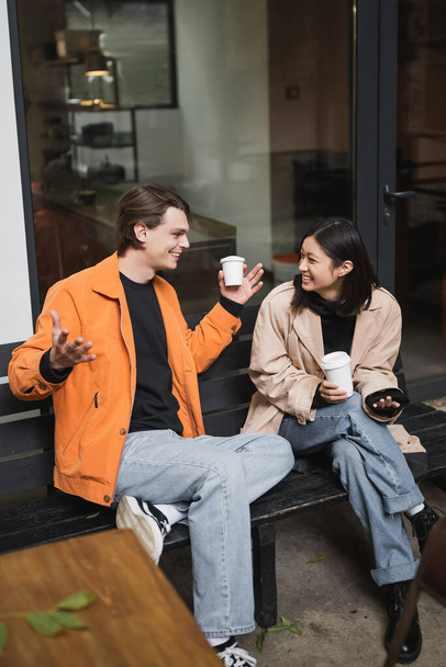 Cheerful asian woman talking to boyfriend with coffee to go on bench near cafe  - 写真・画像