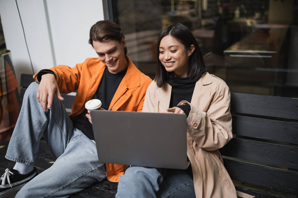 Souriant asiatique femme pointant vers ordinateur portable près de petit ami avec du café pour aller sur banc près de café  - Photo, image