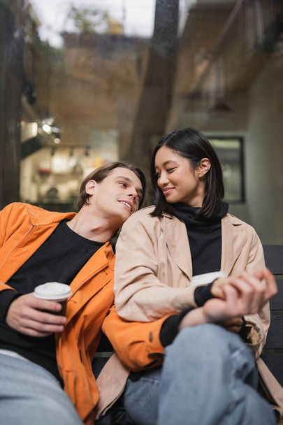 Νεαρό πολυεθνικό ζευγάρι κρατώντας καφέ για να πάει και τα χέρια, ενώ κάθεται στον πάγκο κοντά στο καφέ  - Φωτογραφία, εικόνα