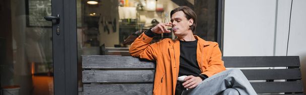 Stijlvolle man met afhaaldrankje en rookdrankje op bankje bij café, spandoek  - Foto, afbeelding