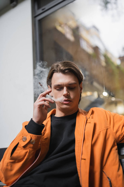 Low angle view of stylish young man smoking cigarette outdoors  - Foto, afbeelding
