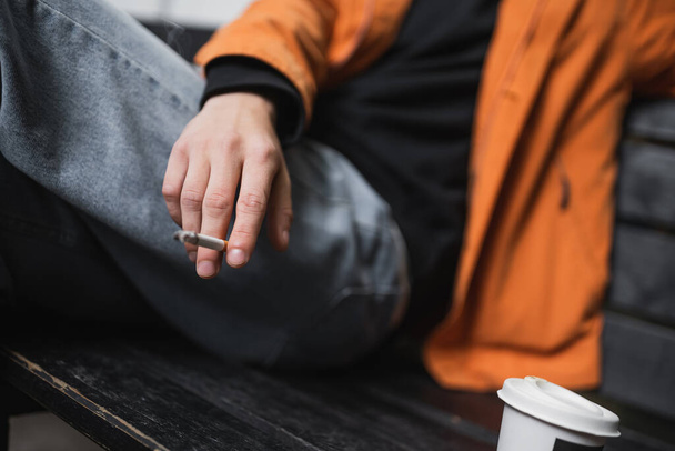 Vista recortada del hombre borroso sosteniendo el cigarrillo cerca del café para ir en el banco al aire libre  - Foto, Imagen