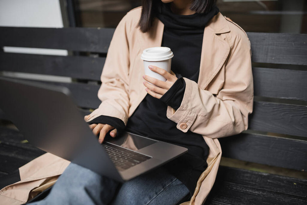 Vue recadrée d'une jeune femme en trench coat tenant une tasse en papier et utilisant un ordinateur portable sur un banc à l'extérieur  - Photo, image