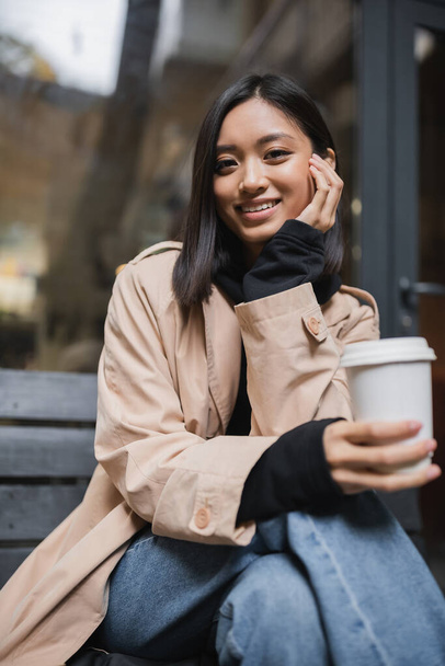 Alegre mujer asiática en gabardina sosteniendo borrosa taza de papel y mirando a la cámara cerca de café  - Foto, Imagen