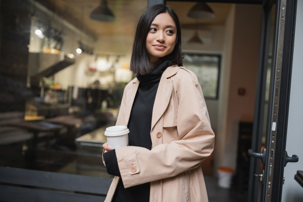 Stylish asian woman holding takeaway coffee near door of cafe outdoors  - Photo, Image
