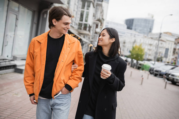 Sonriente pareja multiétnica con taza de papel caminando por la calle urbana  - Foto, Imagen