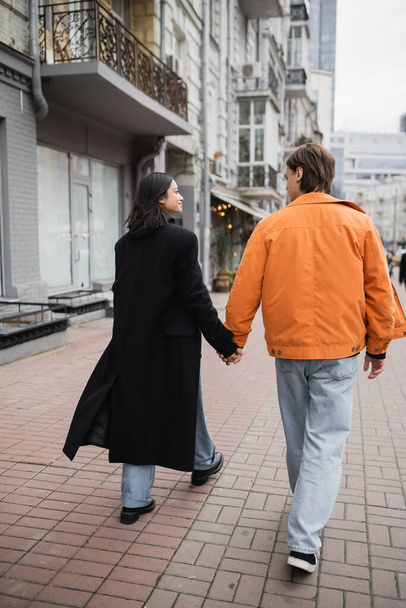 Boční pohled na usmívající se mezirasový pár držící ruce při chůzi na městské ulici  - Fotografie, Obrázek