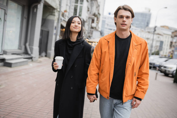Stylish man holding hand of asian girlfriend with coffee to go on urban street  - Photo, Image