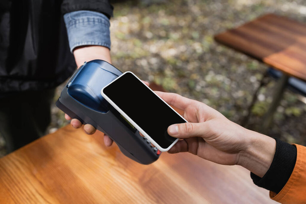 Vue recadrée du jeune homme payant avec smartphone près de la serveuse avec terminal de paiement dans un café extérieur  - Photo, image