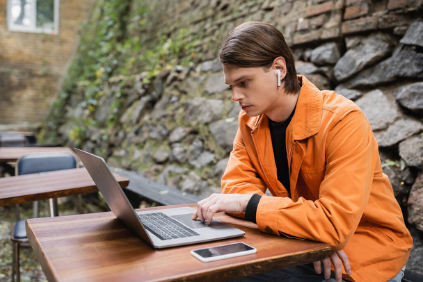 Trendy muž ve sluchátku pomocí notebooku na terase kavárny  - Fotografie, Obrázek