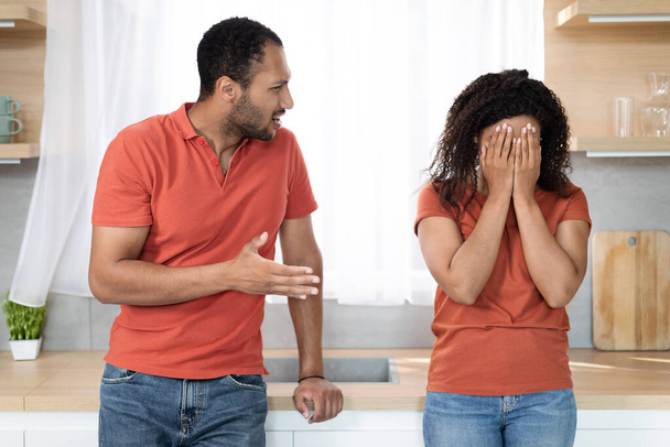 Aggressive angry millennial black husband swears at wife, offended sad woman in red t-shirt crying at quarrel in kitchen interior. Domestic violence, scandal, relationship problems and stress at home - Photo, Image