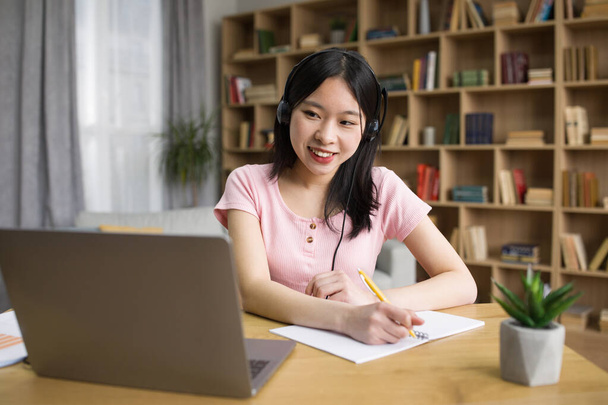 Позитивная азиатская студентка учится перед ноутбуком и делает заметки, носит беспроводные наушники, копирует пространство. Веселая корейская леди посещает вебинар, оставаясь дома во время изоляции - Фото, изображение