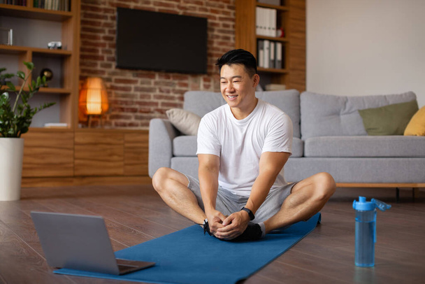 Online yoga praktijk. Rijpe Aziatische man doet vlinder asana op sportmatten, volgende video tutorial op laptop pc, kopieer ruimte. Positieve koreaanse man die thuis traint - Foto, afbeelding