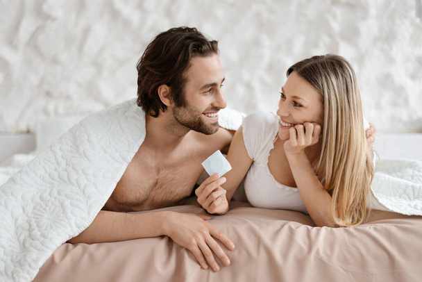 Щаслива любляча пара лежить в ліжку і тримає презерватив, любителі вибирають безпечний секс, використовуючи контрацепцію, копіюють простір. Концепція контролю вагітності та STD
 - Фото, зображення