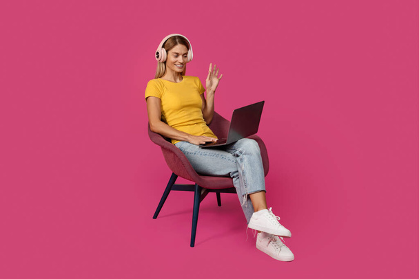 Щаслива зріла біла жінка в повсякденному житті і навушники сидить на кріслі, махаючи рукою на веб-камеру ноутбука, розмовляючи, ізольовані на рожевому фоні, студії. Привіт, привіт, відео дзвінок і зустріч віддалено
 - Фото, зображення