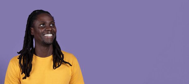 Excited young black guy with long braids looking at copy space for advertisement and smiling on purple studio background, checking nice offer, panorama, closeup shot - Photo, Image