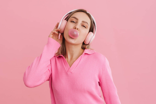 Young cute girl in pink headphones blowing bubble gum over isolated pink background - Foto, immagini