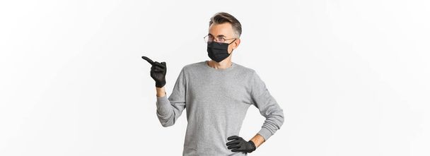 Concept of coronavirus, lifestyle and quarantine. Intrigued middle-aged man in medical mask, gloves and glasses pointing finger right and looking at advertisement, standing over white background. - Photo, Image