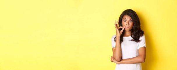 Portrait of young african-american woman making promise to keep secret, seal lips, zipping mouth with fingers, standing over yellow background. - Photo, Image