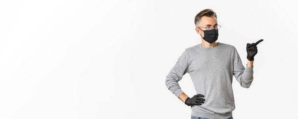 Concept of coronavirus, lifestyle and quarantine. Intrigued middle-aged man in medical mask, gloves and glasses pointing finger right and looking at advertisement, standing over white background. - Photo, Image