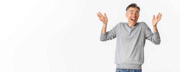 Εικόνα ενός ενθουσιασμένου και ευτυχισμένου μεσήλικα άντρα με γκρίζο κοντό χτένισμα, να σηκώνει τα χέρια ψηλά και να ζητωκραυγάζει, να στέκεται πάνω από λευκό φόντο. - Φωτογραφία, εικόνα
