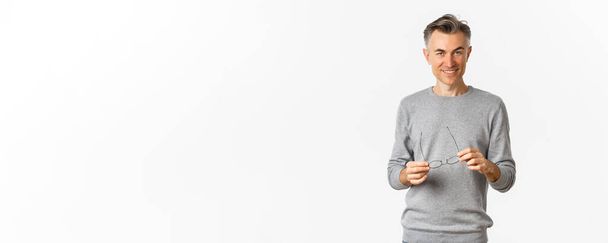 Portrait of handsome and confident man in grey sweater, taking-off glasses and smiling pleased, standing over white background. - Фото, изображение