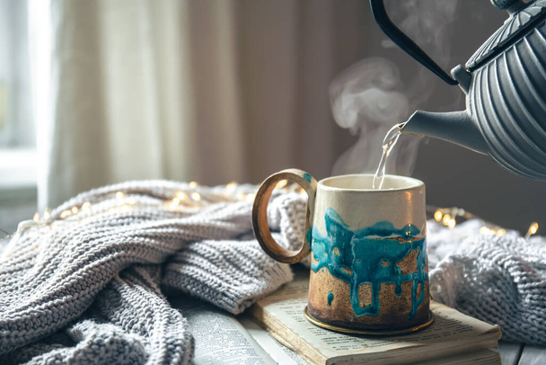 Krásný ručně vyrobený keramický šálek a konvice na rozmazaném pozadí s pleteným prvkem, proces nalévání čaje v pohybu. - Fotografie, Obrázek