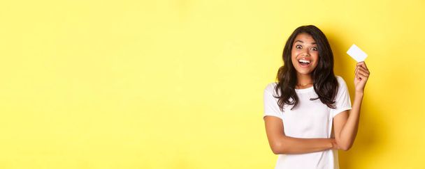 Portret van een verbaasd Afrikaans-Amerikaans meisje, het verhogen van credit card en glimlachen opgewonden, gaan winkelen, staande over gele achtergrond. - Foto, afbeelding