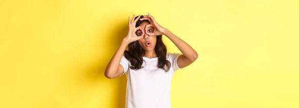 Image of happy and funny african-american girl, making finger glasses and looking through them at something amazing, standing over yellow background. - Photo, Image