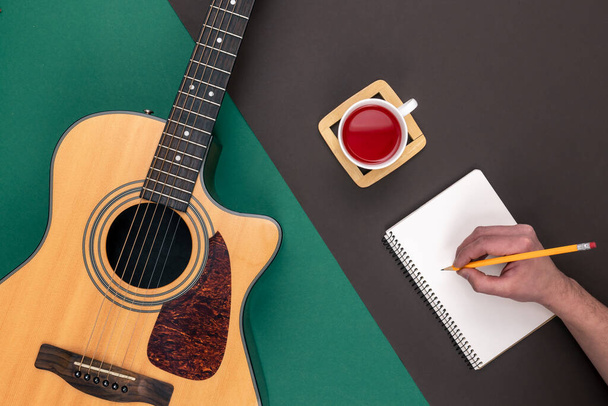 Επίπεδη lay, ακουστική κιθάρα, φλιτζάνι τσάι και σημειωματάριο στο φόντο χρώμα, μουσική κατασκευή και δημιουργικότητα έννοια. - Φωτογραφία, εικόνα