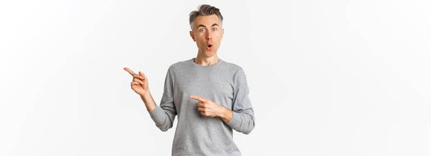 Amazed handsome guy in grey sweater, pointing fingers right and looking curious, askign question about product, standing over white background. - Foto, imagen