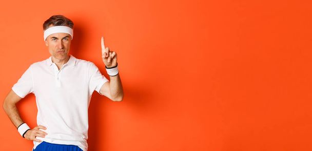 Portrait of serious middle-aged male athlete, wearing sportswear, pointing finger up and showing logo about sports and gym, standing over orange background. - Photo, Image