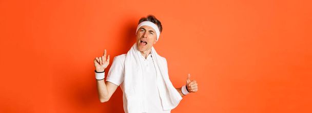 Image of happy and carefree middle-aged fitness guy, dancing after workout, holding towel over neck, posing against orange background. - Photo, Image