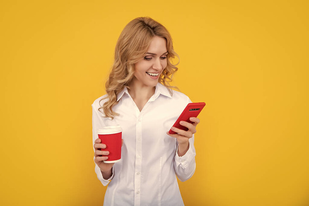 χαρούμενη ξανθιά γυναίκα με φλιτζάνι καφέ κουβεντιάζοντας στο τηλέφωνο σε κίτρινο φόντο. - Φωτογραφία, εικόνα