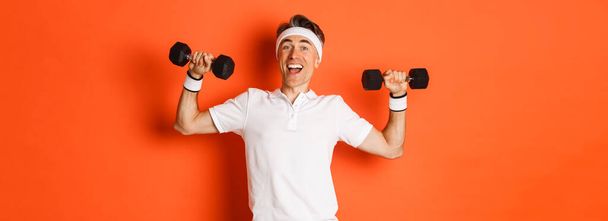 Conceito de treino, ginásio e estilo de vida. Imagem de atleta masculino de meia-idade saudável e forte, fazendo exercícios esportivos com halteres e sorrindo, em pé sobre fundo laranja. - Foto, Imagem
