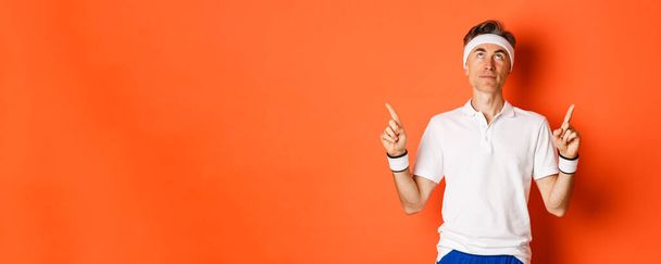 Portrait of handsome middle-aged male athlete, wearing sports clothing, looking and pointing fingers up at promo banner, standing against orange background. - Foto, imagen